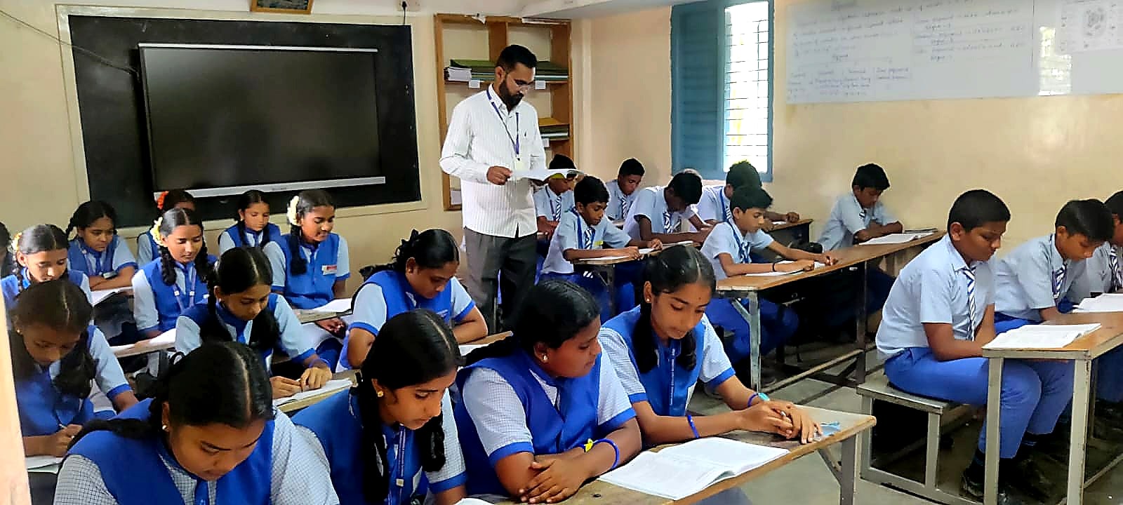 A senior classroom in session at Manjushree — students in blue uniform writing at their desks with the teacher walking through